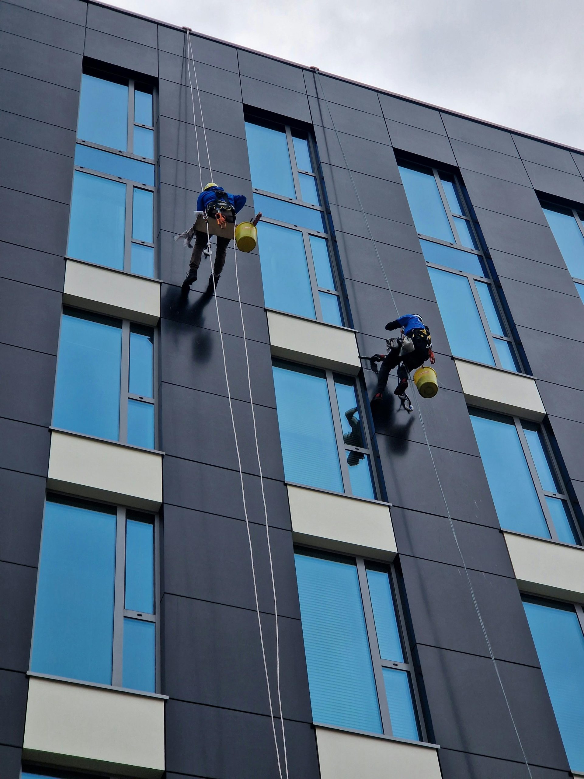 Zwei Arbeiter reinigen mit Sicherheitsseilen in der Höhe die Fassade eines modernen Gebäudes.