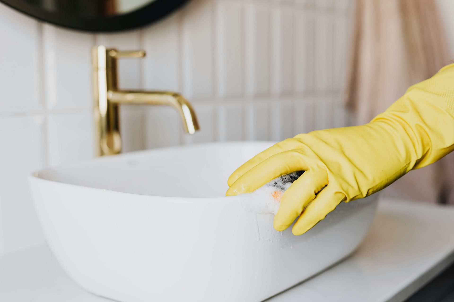 Eine gesichtslose Person in gelben Latexhandschuhen reinigt die Oberfläche eines weißen, eleganten Waschbeckens in einem modernen Badezimmer.